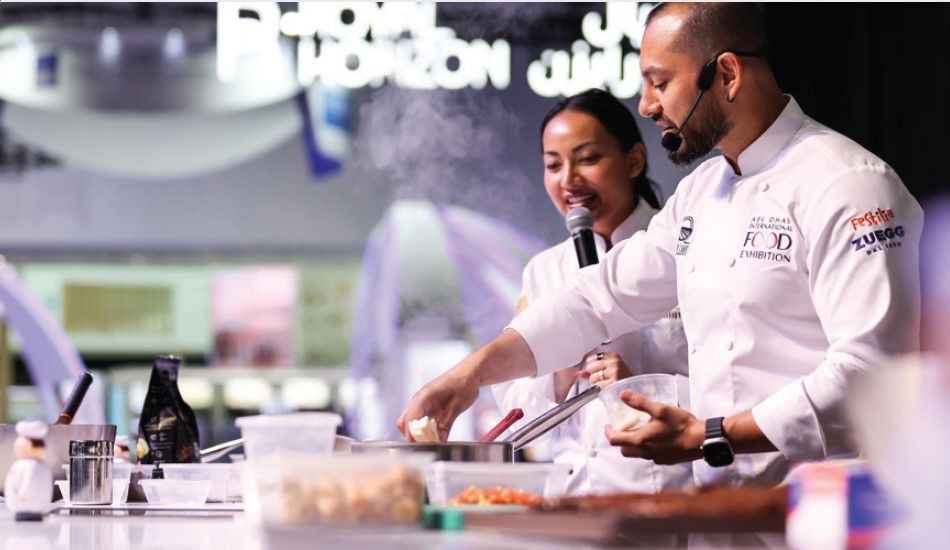 Chef demonstrating cooking techniques at a food exhibition, with various cooking utensils and ingredients on the counter.